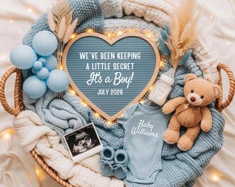 May include: A wicker basket containing baby essentials: a blue onesie, booties, a teddy bear, and a heart-shaped sign saying "It's a Boy!" and "July 2026." The display also features blue balloons and a baby bottle.