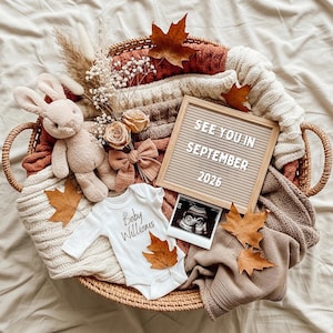 Puede incluir: Una cesta de mimbre llena de un conejito de peluche, ropa de bebé y hojas de otoño. Un tablero de letras dice "SEE YOU IN SEPTEMBER 2026". Una imagen de ultrasonido y un lazo también están en la cesta, anunciando un embarazo.