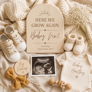 Puede incluir: Un letrero de madera arqueado con el texto Here We Grow Again Baby No.2 Arriving September 2027, un par de zapatillas beige, patucos de punto blancos, un body acanalado con Baby Hartley, un disco de madera Big Sibling, un lazo mostaza y una ecografía.