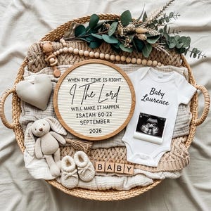 Peut inclure: Un panier en osier contient des articles pour bébé, dont un body blanc avec "Baby Laurence" imprimé, une échographie et un panneau indiquant "When the time is right, I The Lord will make it happen." D'autres articles incluent un lapin en peluche, des chaussures et des blocs de bois.