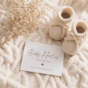 May include: A pair of cream-colored knitted baby booties with tie closures next to a white announcement card that reads Baby Hartley IS ON THE WAY with a heart icon and ARRIVING DECEMBER 202X, all resting on a textured cream cable knit fabric.