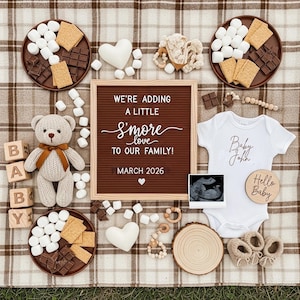 Könnte beinhalten: Eine Babyankündigung auf einer karierten Decke. Es gibt eine Buchstaben-Tafel mit dem Text "We're adding a little S'more love to our family! March 2026". Dazu ein Teddybär, Babykleidung und Zutaten für S'mores wie Schokolade und Marshmallows.