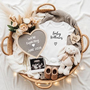 Może przedstawiać: Wiklinowy kosz zawiera elementy ogłoszenia o dziecku. Białe body z napisem "Baby Williams" i sercem, tablica w kształcie serca z napisem "We're Expecting June 2026", pluszowy królik, buciki dziecięce i zdjęcie USG.