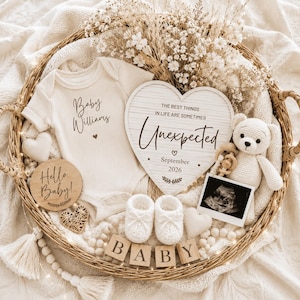 May include: A cream-colored ribbed baby bodysuit with the text Baby Williams, a heart-shaped sign reading The best things in life are sometimes Unexpected September 2026, a crochet teddy bear, white booties, wooden blocks spelling BABY, and an ultrasound.