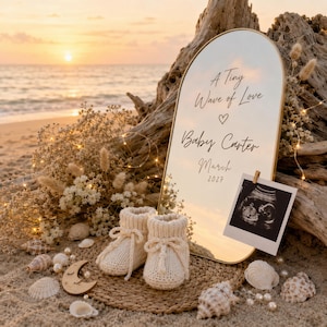 Puede incluir: Un espejo arqueado con borde dorado que presenta el texto A Tiny Wave of Love, Baby Carter y March 2027. Una ecografía en blanco y negro está sujeta al espejo. Un par de patucos de punto color crema descansan sobre una esterilla con conchas.