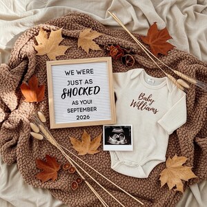 May include: A baby announcement with a letter board saying "WE WERE JUST AS SHOCKED AS YOU! SEPTEMBER 2026", a cream-coloured babygrow with "Baby Williams", and an ultrasound scan. The scene is decorated with autumn leaves and other seasonal elements.