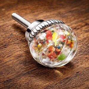 May include: A clear, round cabinet knob with a silver base and screw. The knob's glass sphere is filled with colorful, small pieces of red, yellow, blue, and green materials. The knob is set against a wooden surface.
