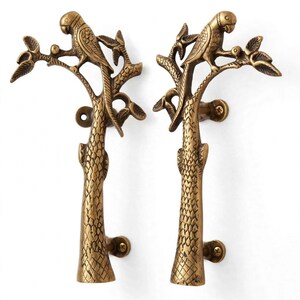May include: A pair of brass door handles featuring a textured tree trunk design with a parrot perched on a branch at the top. Each handle includes two mounting points with visible screws for attachment to a door surface, finished in a metallic gold.
