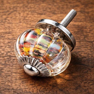 Könnte beinhalten: Ein klarer Glasschrankknopf mit einem farbenfrohen, geschichteten Innenraum. Der Knopf hat eine silberne Metallbasis und einen Gewindestift zur Installation. Der farbenfrohe Innenraum umfasst Gelb-, Rot-, Grün- und Blautöne. Der Knopf befindet sich auf einer Holzoberfläche.