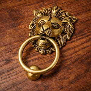 Puede incluir: Un aldaba de puerta con cabeza de león de latón con un asa circular y una pequeña bola en la base. La cabeza del león está detallada con una melena y rasgos faciales. La aldaba está montada sobre una superficie de madera.