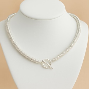 Può includere: Una collana argentata con chiusura a toggle, esposta su un busto di manichino bianco. La collana presenta un design strutturato e arrotolato. La chiusura è una semplice chiusura a barra e anello. Lo sfondo è di colore neutro.