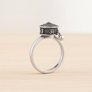Puede incluir: Un anillo de plata con un diseño de rueda de oración. El anillo tiene una banda pulida y una parte superior texturizada con símbolos grabados. Una pequeña cuenta cuelga de la rueda. El anillo es una pieza de joyería espiritual.