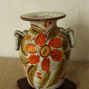 Può includere: Un vaso in ceramica dipinto a mano con un motivo floreale. Il vaso presenta uno sfondo grigio maculato con fiori arancioni e gialli e foglie verdi. Ha due manici e poggia su una piccola base quadrata in legno marrone scuro.