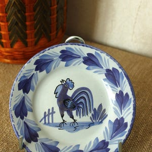 Könnte beinhalten: Dekorative Keramikplatte mit einem Hahn-Design in Blau und Weiß. Die Platte hat einen blauen Blumenrand und wird auf einem Metallständer präsentiert. Ein Weidenkorb befindet sich im Hintergrund.