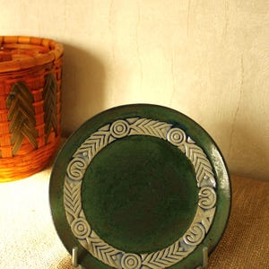 Puede incluir: Un plato de cerámica verde oscuro con un borde decorativo blanco y azul. El plato se exhibe sobre un soporte metálico. Una cesta tejida es visible en el fondo.