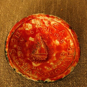 May include: A round, red ceramic object with embossed text and a sailboat design. The text includes the date "6 Avril 1968". The object has a dark blue rim and is set against a textured, brown background.