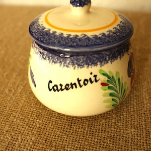 Puede incluir: Un pequeño tarro de cerámica redondo con tapa. El tarro es de color crema con un borde y una tapa con dibujos azules y blancos. La palabra "Carentoir" está escrita en la parte delantera. También hay detalles florales verdes y rojos.