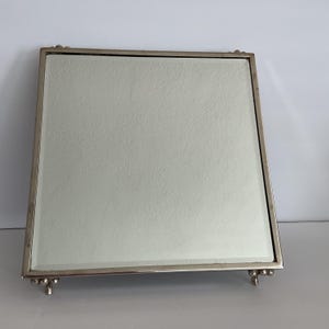 May include: A square mirror tray with a polished silver-toned metal frame and a reflective glass surface. The tray is elevated by four small, decorative ball-shaped feet at each corner, providing a stable base for this reflective display piece.
