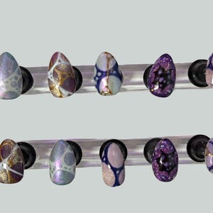 May include: A set of ten artificial nails with various designs. The nails are oval-shaped and feature a range of colors, including purple, gold, blue, and white. Each nail has a unique pattern, such as swirls, dots, and abstract shapes. They are displayed on clear stands.