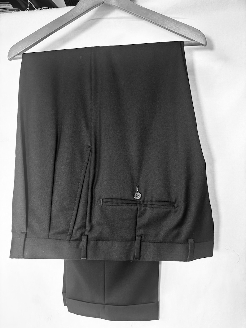 May include: Black dress pants hanging on a wooden hanger. The pants have a buttoned back pocket and belt loops. The bottom of the pants are cuffed.