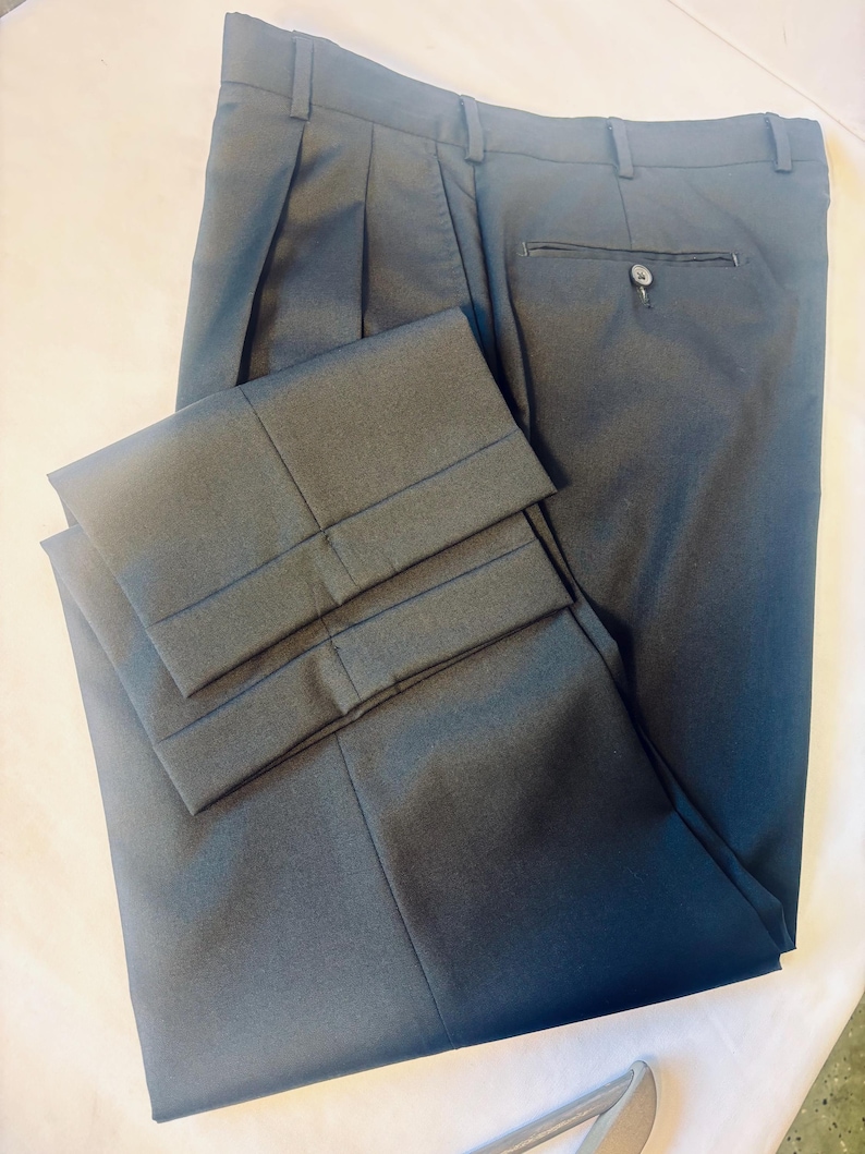 May include: Dark gray pleated dress pants. The pants have a button closure, belt loops, and a back pocket. The cuffs are folded up. The pants are made of a smooth fabric and are neatly folded.