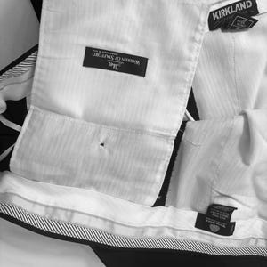 May include: Close-up of black dress pants with white lining and labels. The labels read "Warren of Stafford" and "Kirkland". The pants have a striped waistband and are made of wool.