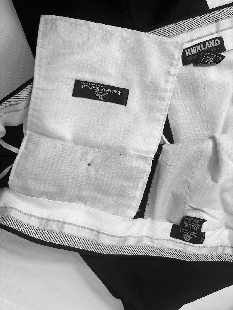 May include: Close-up of black dress pants with white lining and labels. The labels read "Warren of Stafford" and "Kirkland". The pants have a striped waistband and are made of wool.