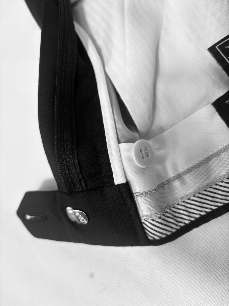 May include: Close-up of a black and white suit pant. The image shows the button closure, zipper, and interior lining. The white lining has a subtle striped pattern. The black fabric is smooth and tailored.