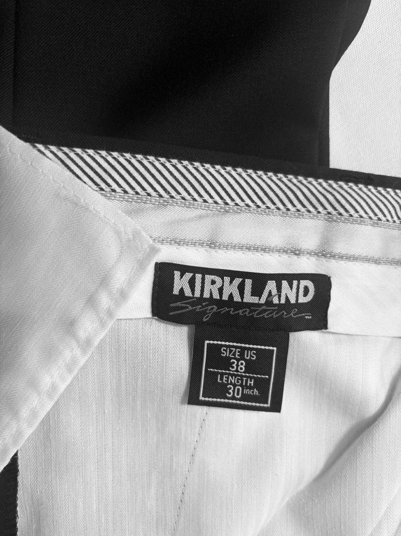 May include: Close-up of a black and white label from Kirkland Signature pants. The label indicates a US size 38 with a 30-inch length. The pants are black with a white interior and a striped waistband.