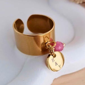 Puede incluir: Un anillo ajustable de color dorado con una pequeña cuenta rosa y un dije redondo. El dije está grabado con las letras "E & Y". El anillo está sobre una superficie blanca.