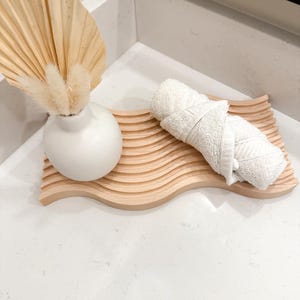May include: A light-coloured wooden tray with a wavy design holds a rolled white towel. A small white vase with dried flowers and a large dried palm leaf are also on the countertop. The items are arranged for bathroom decor.