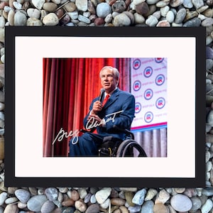 Puede incluir: Fotografía enmarcada de un hombre con traje azul y corbata roja, sentado en una silla de ruedas, hablando por un micrófono. El fondo muestra una cortina roja y logotipos. La fotografía incluye una firma manuscrita.
