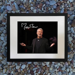 Può includere: Fotografia incorniciata di un uomo in abito nero, con in mano un piccolo dispositivo, con le braccia aperte. La fotografia è firmata con il nome "Michael S." in grafia bianca. Lo sfondo è scuro.