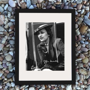 以下が含まれることがあります： 帽子と手袋を着用した女性の白黒写真。女性は座って横を見ています。写真は右下に筆記体で署名されています。