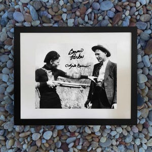 May include: Black and white framed photograph featuring Bonnie Parker and Clyde Barrow. The image shows Bonnie Parker holding a shotgun, with Clyde Barrow reaching out. The signatures "Bonnie Parker" and "Clyde Barrow" are visible.