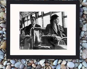 May include: Black and white framed print featuring Rosa Parks on a bus. The image shows two people seated on a bus, with Rosa Parks' signature on the seat. The print is set against a background of small, colorful stones.