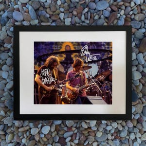 Może przedstawiać: Oprawiona fotografia przedstawiająca dwóch muzyków grających na gitarach na scenie. Na zdjęciu widnieją podpisy białym tuszem. Tło to kolorowy wzór tie-dye. Rama jest czarna, a zdjęcie na białym passe-partout.