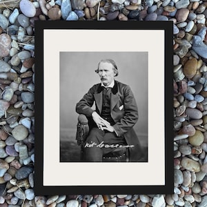 May include: Black and white framed portrait of a man seated in a chair. The man is wearing a dark suit and has a mustache. The image is signed in cursive at the bottom. The frame is black.