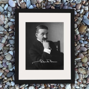 May include: Black and white framed portrait of a man with a mustache, wearing a suit and tie. The man is seated and has his hand near his face. The signature "Frank Baum" is visible at the bottom of the image.