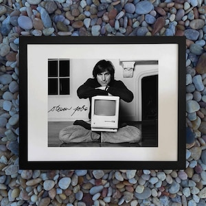 Puede incluir: Fotografía enmarcada en blanco y negro con Steve Jobs y un ordenador Macintosh vintage. La fotografía incluye la firma manuscrita "steven jobs". La imagen está situada sobre un fondo de una ventana y una chimenea.