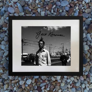 May include: Black and white framed print featuring a man standing on a city street. The print includes the handwritten signature "Jack Kerouac" in the upper right corner. The image is set against a backdrop of a cloudy sky and urban landscape.