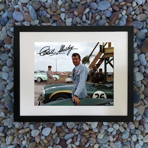 Puede incluir: Impresión enmarcada con la firma de Carroll Shelby sobre una fotografía de él con un coche de carreras verde. La foto muestra a Shelby con un mono azul claro, apoyado en el coche. Otros coches y personas antiguas están en el fondo.