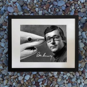 May include: Black and white framed photograph featuring a man with glasses and a shark. The shark's open mouth is near the man's face. The photograph is signed "Peter Benchley".