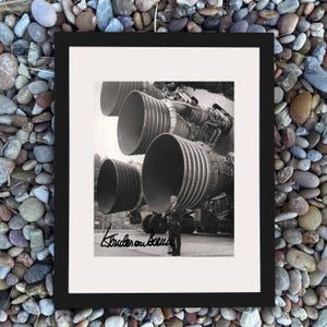 Puede incluir: Impresión en blanco y negro enmarcada que presenta grandes motores de cohetes. Un hombre con traje está de pie cerca de los motores. La impresión está firmada en la esquina inferior izquierda. El marco es negro.