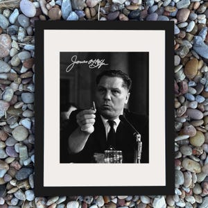 Puede incluir: Fotografía enmarcada en blanco y negro que muestra a un hombre con traje sosteniendo un bolígrafo. La fotografía tiene una firma en la esquina superior izquierda. El hombre está sentado en una mesa con un micrófono.