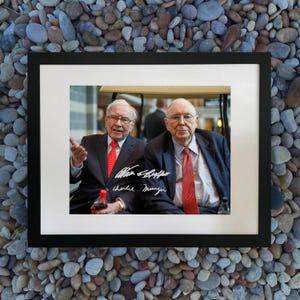 May include: Framed print showing two men in suits and red ties, one holding a bottle. The print includes signatures. The frame is black, and the print is matted in white. The background is a pebble surface.
