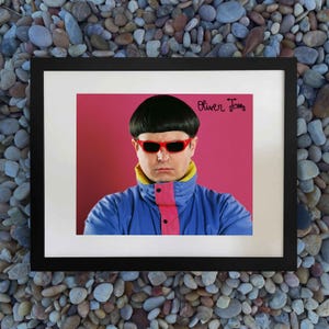 May include: A framed print featuring a person with a bowl cut hairstyle, wearing red-rimmed sunglasses and a blue jacket with a pink center panel. The background is solid pink, and the text Oliver Tree is printed in black with a small scooter icon.