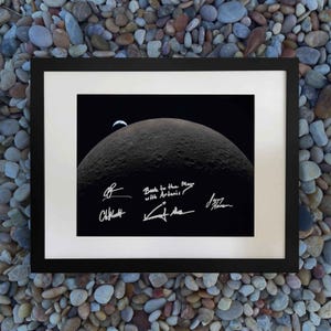 Pode incluir: Impressão emoldurada com uma imagem detalhada da superfície lunar com um corpo celeste menor ao fundo. A impressão inclui assinaturas e o texto "Back to the Moon with Artemis!" em branco sobre um fundo preto.