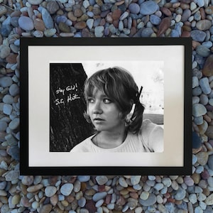 May include: Black and white framed photograph of a young woman with her hair in a ponytail. The photograph has the text "stay gold! S.E. Hint" written on the left side. The frame is black.