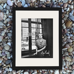 Pode incluir: Fotografia emoldurada em preto e branco de uma mulher sentada perto de uma janela, com a assinatura cursiva "Ayn Rand". A janela tem vista para uma rua da cidade com edifícios altos.